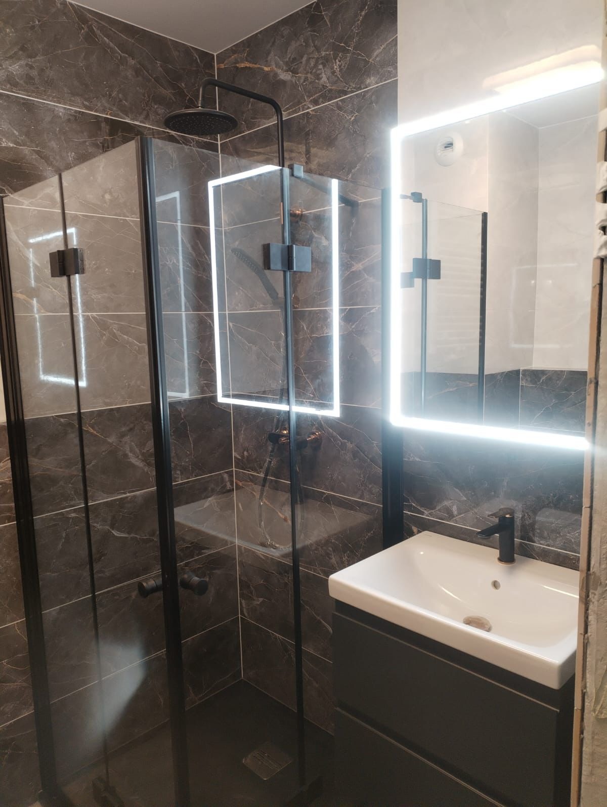 Nowoczesna łazienka z kabiną prysznicową typu walk-in z czarnymi profilami, deszczownicą i marmurowymi płytkami, umywalka nablatowa z szafką i lustrem z podświetleniem LED.