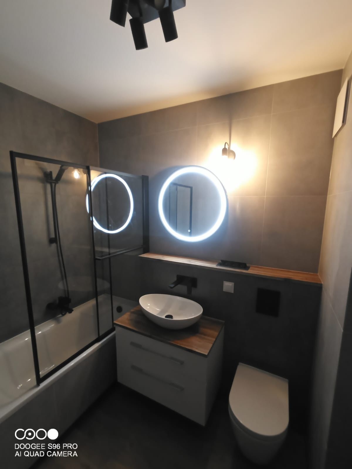 Nowoczesna łazienka z szarymi płytkami, okrągłymi lustrami z podświetleniem LED, białą umywalką nablatową na szafce z drewnianym blatem i kabiną prysznicową z czarnymi ramami.