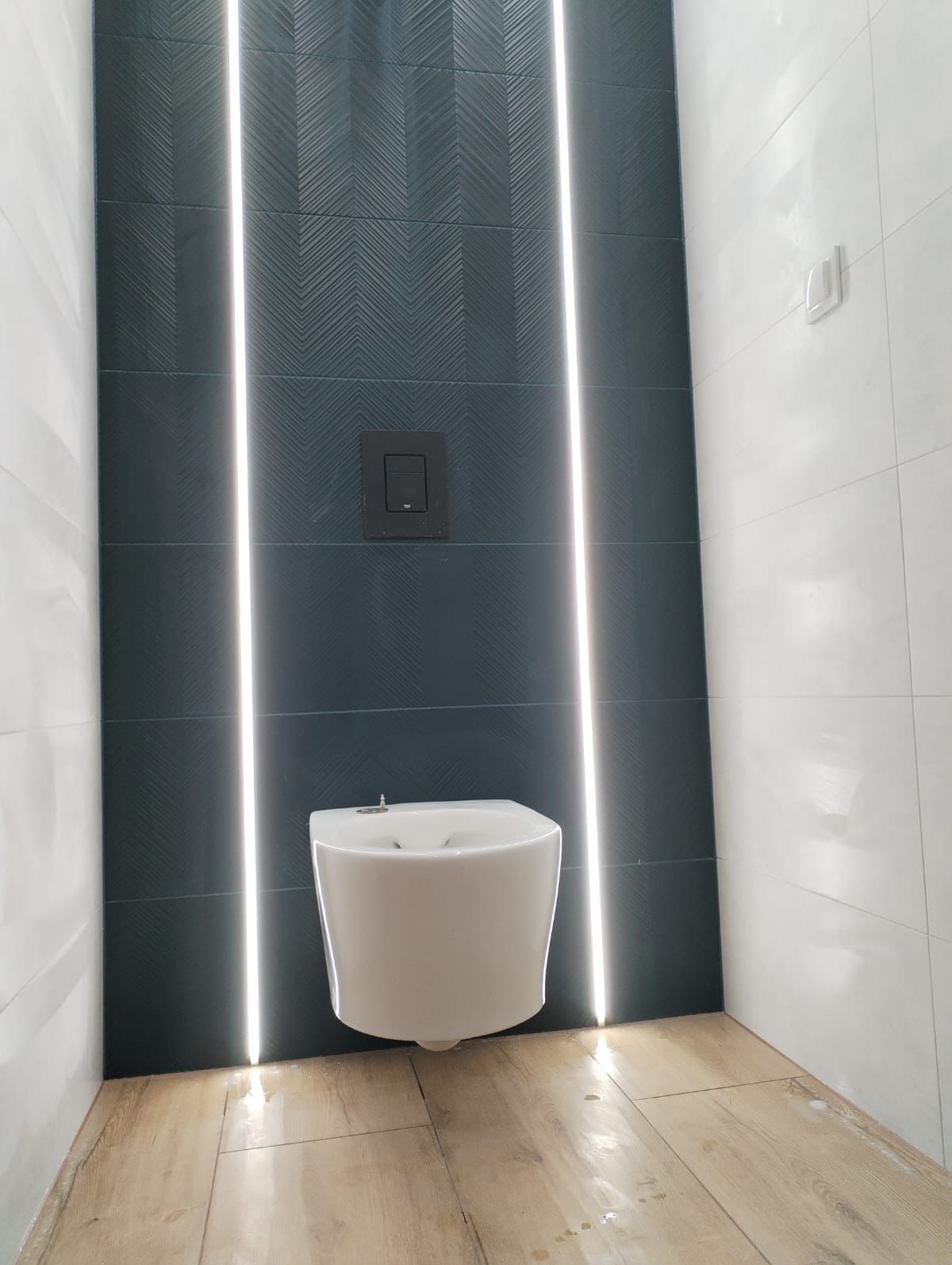 Nowoczesna toaleta podwieszana na ścianie z ciemnymi płytkami w jodełkę i białymi płytkami po bokach, oświetlona pionowymi pasami LED.