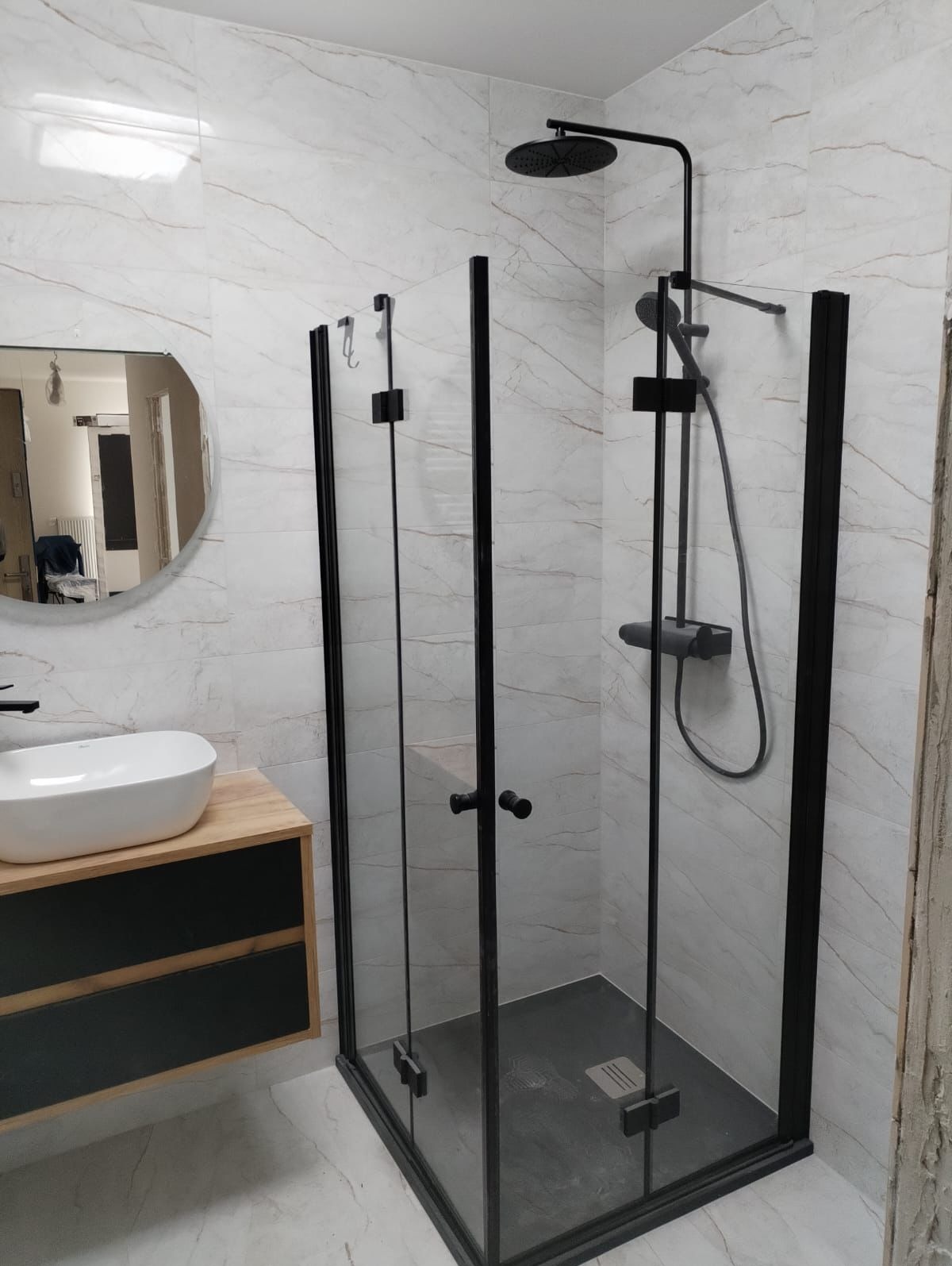 Nowoczesna łazienka z kabiną prysznicową typu walk-in z czarnymi profilami, białą umywalką nablatową na drewnianej szafce i marmurowymi płytkami na ścianach.