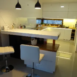 Nowoczesna, jasna kuchnia z białymi, błyszczącymi frontami, drewnianym blatem i barkiem, oświetlona lampami wiszącymi, z dwoma białymi hokerami i elementami dekoracyjnymi.