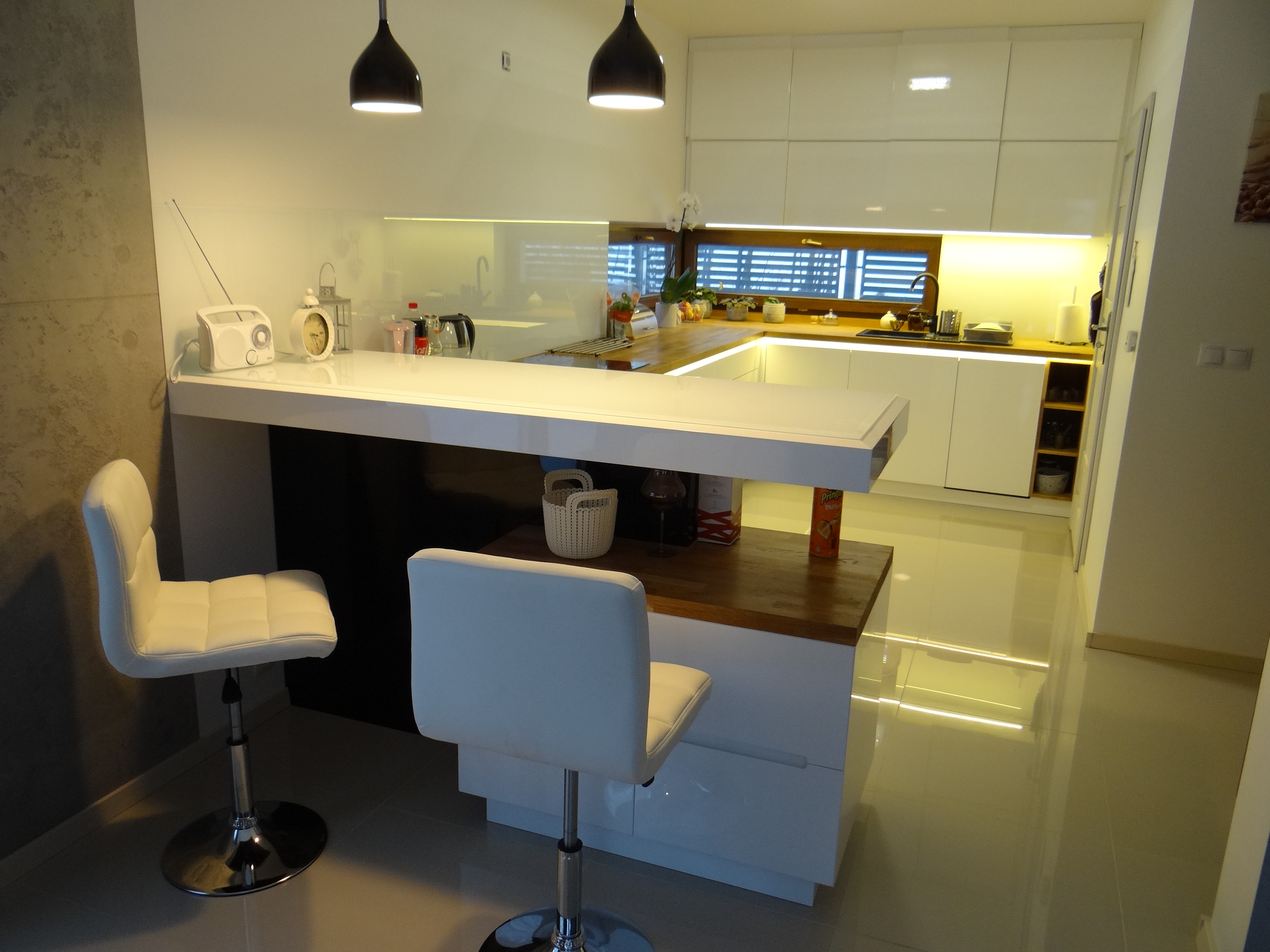 Nowoczesna, jasna kuchnia z białymi, błyszczącymi frontami, drewnianym blatem i barkiem, oświetlona lampami wiszącymi, z dwoma białymi hokerami i elementami dekoracyjnymi.