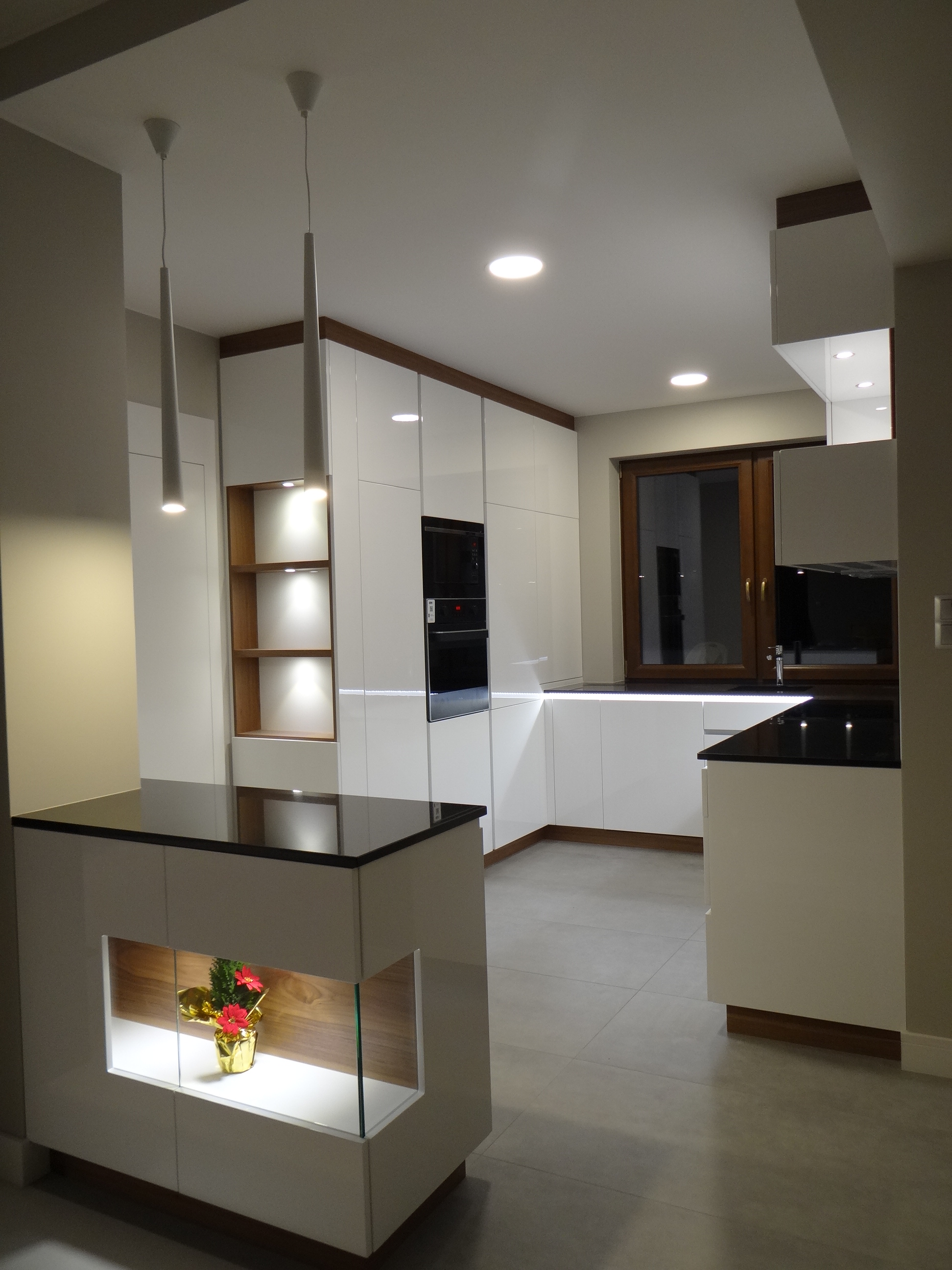 Nowoczesna kuchnia z białymi, lakierowanymi frontami, drewnianymi akcentami i wyspą z podświetlaną wnęką na dekorację.