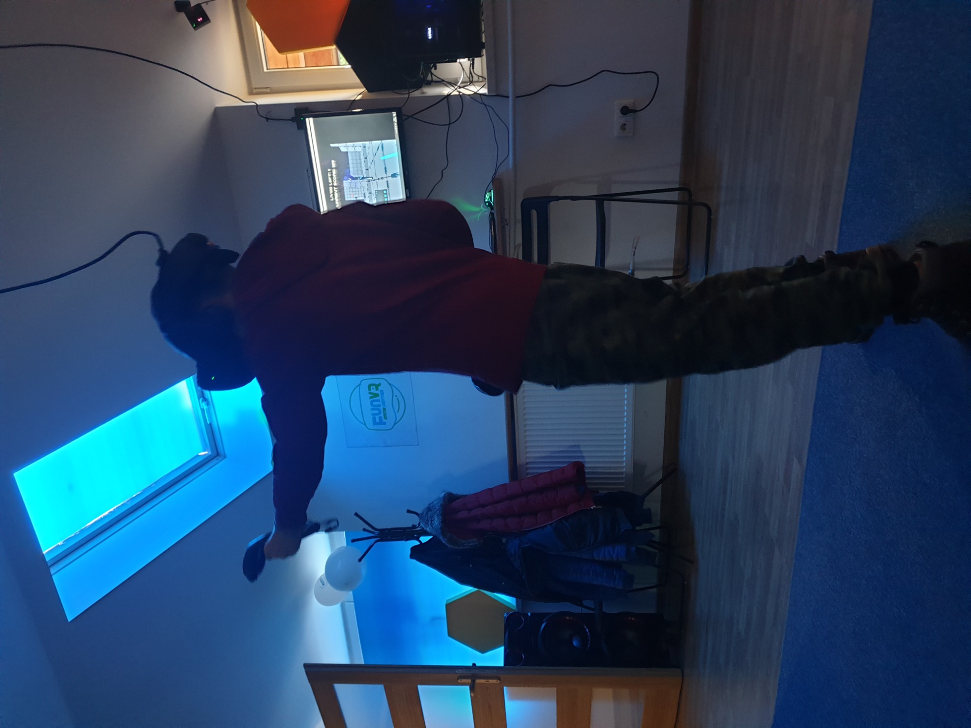 Osoba w czerwonej bluzie i goglach VR gra w wirtualną rzeczywistość w pokoju z niebieskim oświetleniem i oknem dachowym.