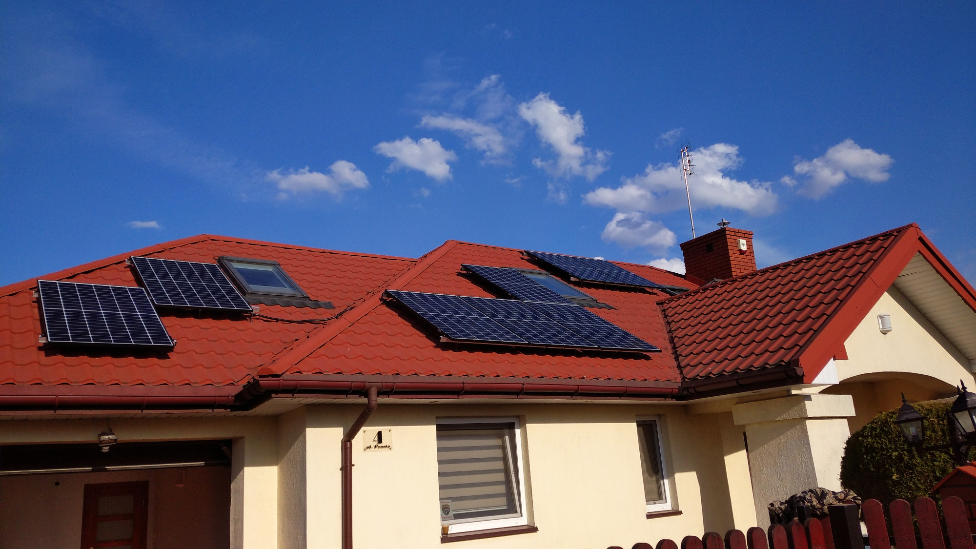 Panele słoneczne na dachu domu z czerwoną dachówką, widoczne okna dachowe i komin z anteną, błękitne niebo z chmurami w tle.