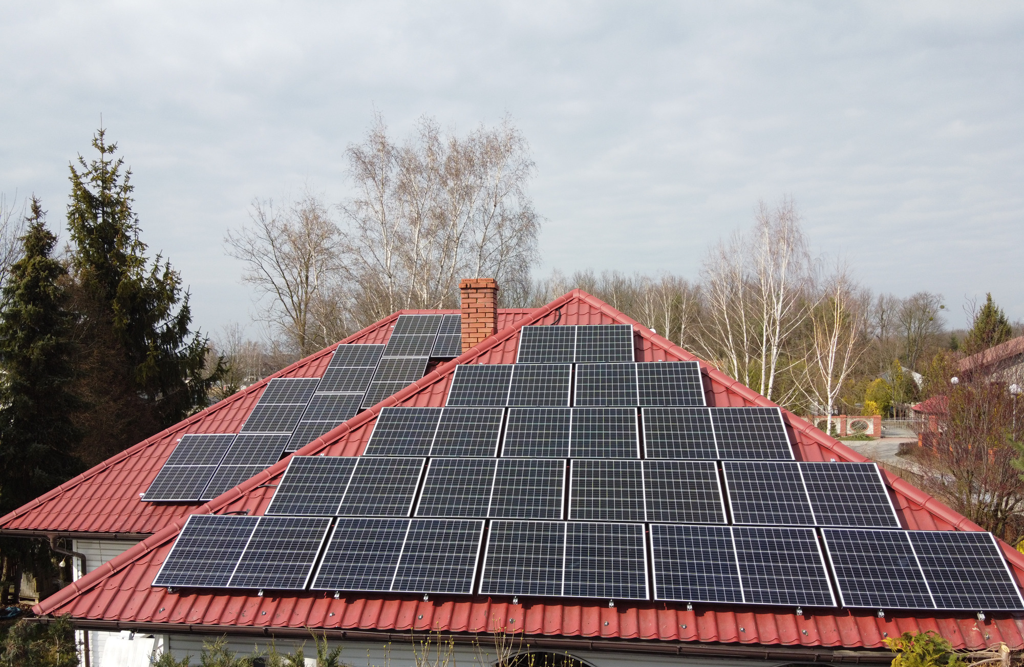 Czerwony dach domu z ceglanym kominem pokryty panelami słonecznymi, widok z dołu w pochmurny dzień.