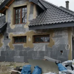 Surowa elewacja domu w trakcie budowy, widoczne szare bloczki, częściowo otynkowane okolice okien, czekające na dalsze prace wykończeniowe, z fragmentami niezagospodarowanego terenu wokół.