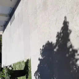 Jasne, kwadratowe płyty tarasowe ułożone na zewnątrz budynku, z widocznym cieniem drzewa i fragmentem zieleni oraz elewacji z oknami zabezpieczonymi folią.