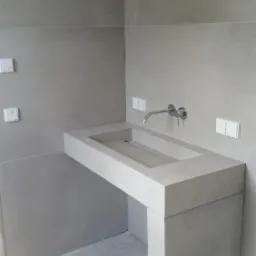 Nowoczesna, minimalistyczna łazienka z umywalką zintegrowaną z blatem i nogą z szarego betonu architektonicznego, chromowana bateria ścienna, szare płytki na ścianach, widoczne gniazdka elektryczne...