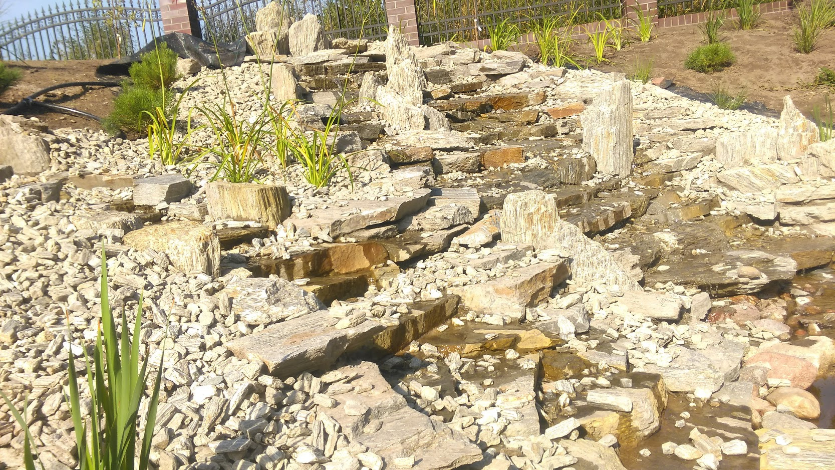 Kaskadowy strumień z płaskich kamieni i drobnego grysu, z roślinnością w tle, tworzący naturalny element dekoracyjny ogrodu.