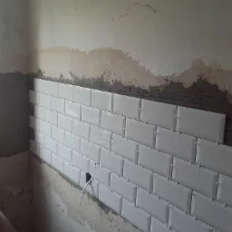 Ściana w trakcie układania białych, prostokątnych płytek typu metro, widoczne krzyżyki dystansowe i surowy stan ścian z wystającymi przewodami elektrycznymi.