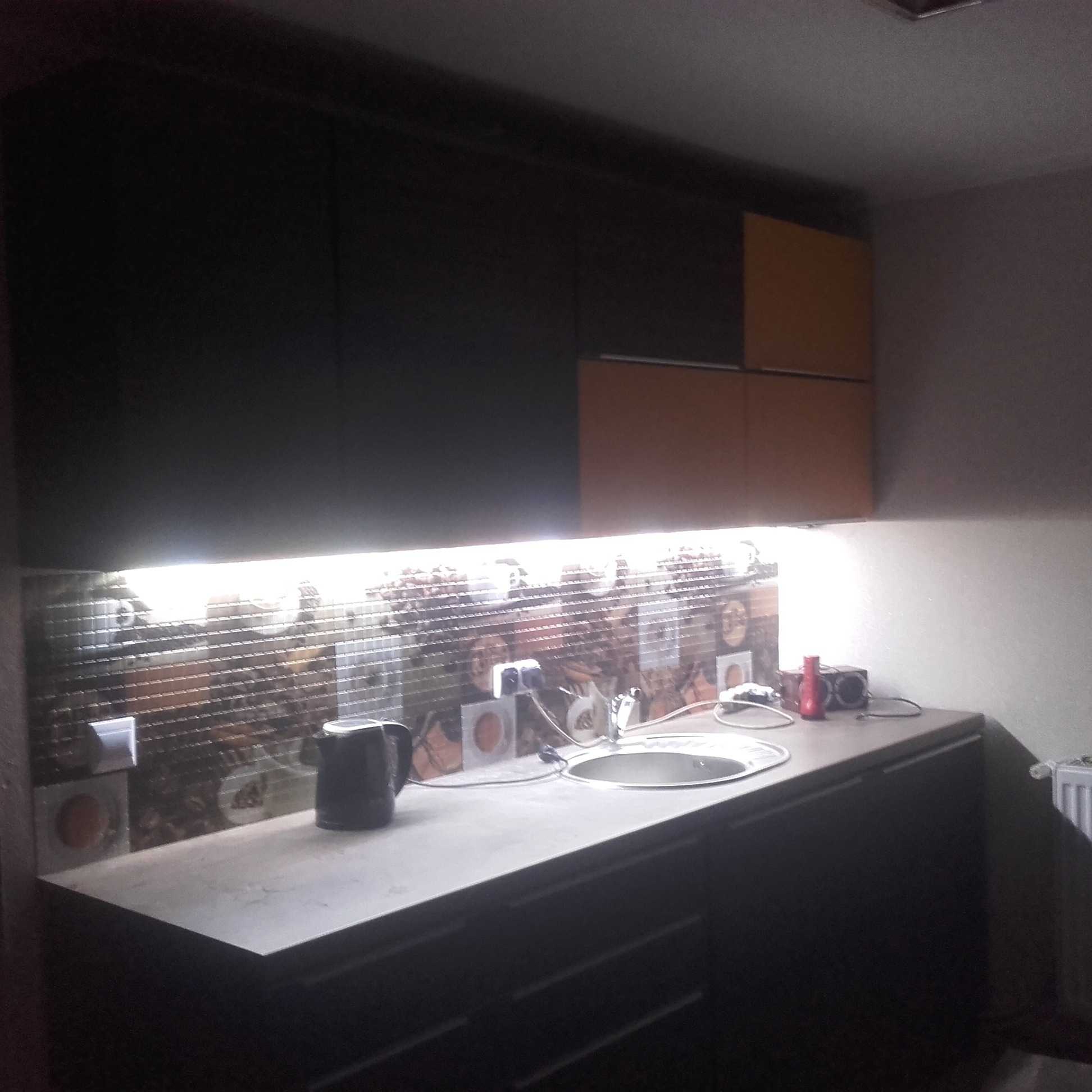 Nowoczesna kuchnia z ciemnymi szafkami, jasnym blatem i mozaiką na ścianie podświetloną ledami, widoczny czajnik elektryczny i zlewozmywak.