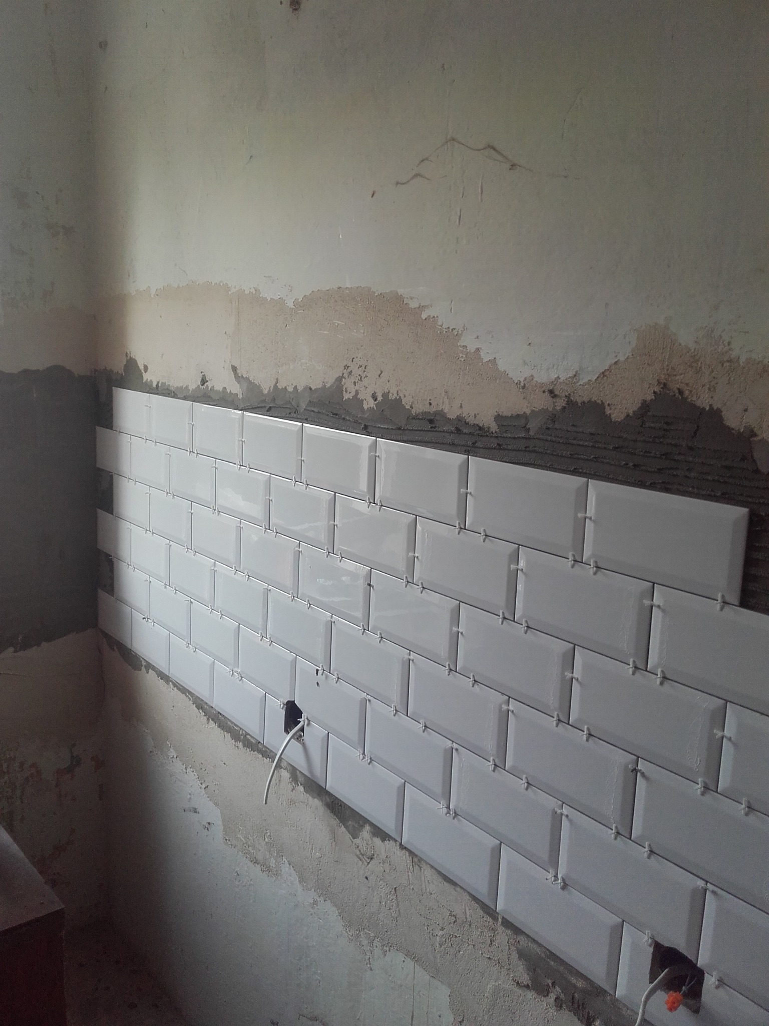 Ściana w trakcie układania białych, prostokątnych płytek typu metro, widoczne krzyżyki dystansowe i surowy stan ścian z wystającymi przewodami elektrycznymi.