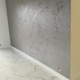 Ściana pokryta dekoracyjną szarą strukturą, widoczne gniazdka elektryczne, biała listwa przypodłogowa i marmurowe płytki podłogowe.