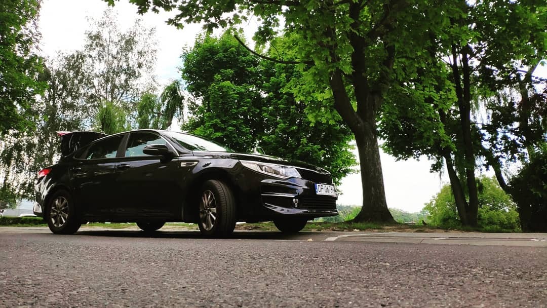 Czarny samochód osobowy z otwartym bagażnikiem zaparkowany na poboczu drogi w otoczeniu zieleni drzew.