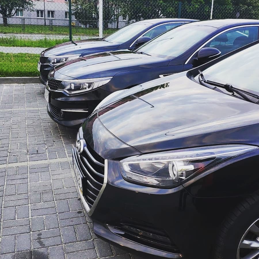 Trzy czarne samochody osobowe zaparkowane w rzędzie na brukowanym parkingu, widoczne przednie reflektory i maski, budynek w tle.