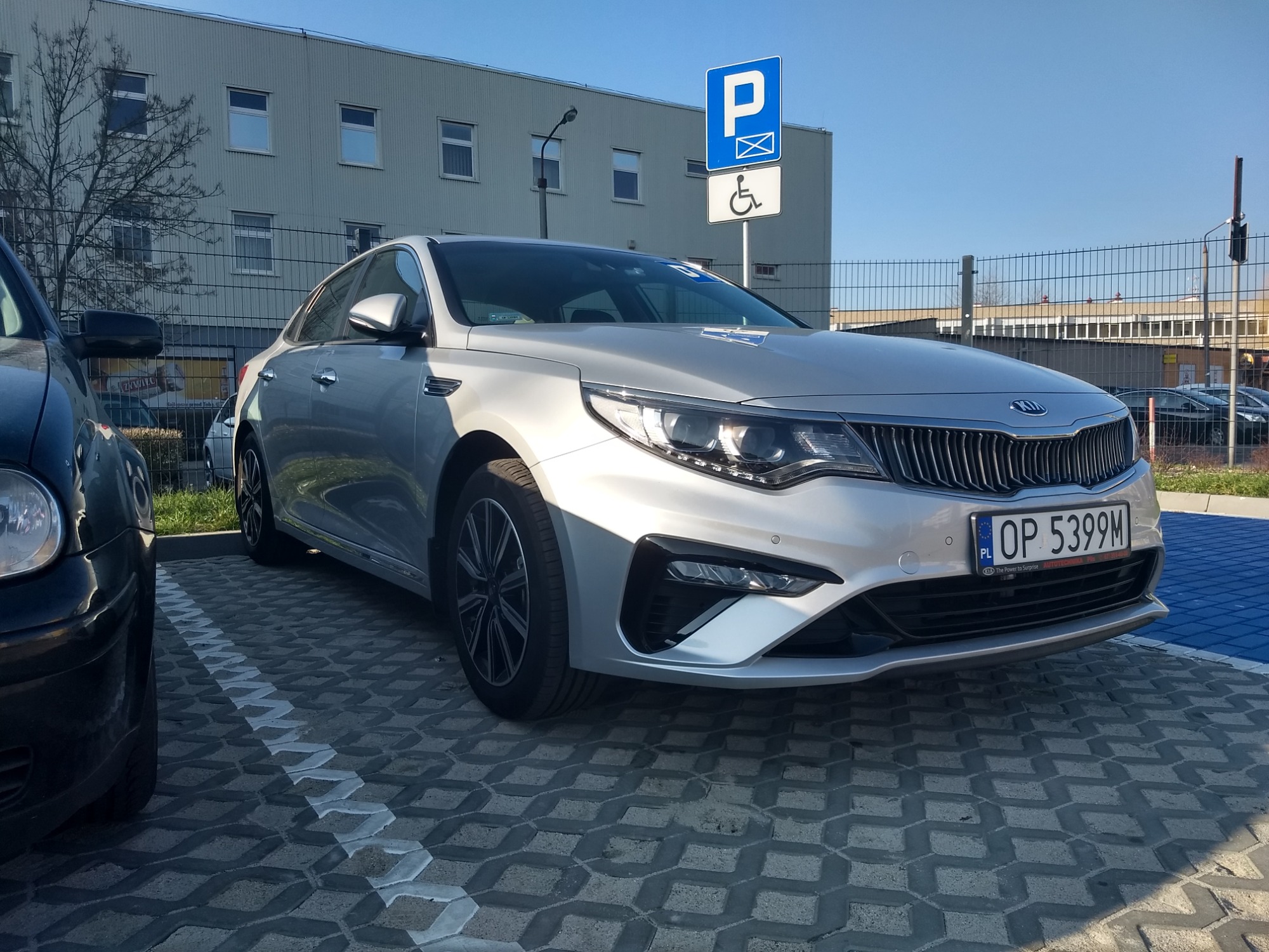 Srebrny samochód osobowy Kia Optima zaparkowany na miejscu dla osób niepełnosprawnych, widoczny znak pionowy z symbolem wózka inwalidzkiego.