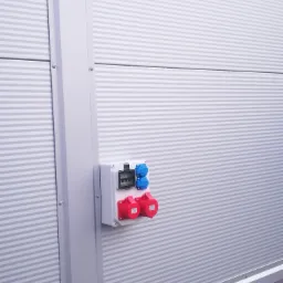 Zewnętrzna skrzynka elektryczna z gniazdami siłowymi (czerwone i niebieskie) zamontowana na szarej, blaszanej ścianie hali.