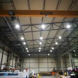 Wnętrze hali produkcyjnej z suwnicą BUDTOR pod sufitem, oświetleniem LED, obrabianymi elementami, wózkami widłowymi i pracownikami w tle.