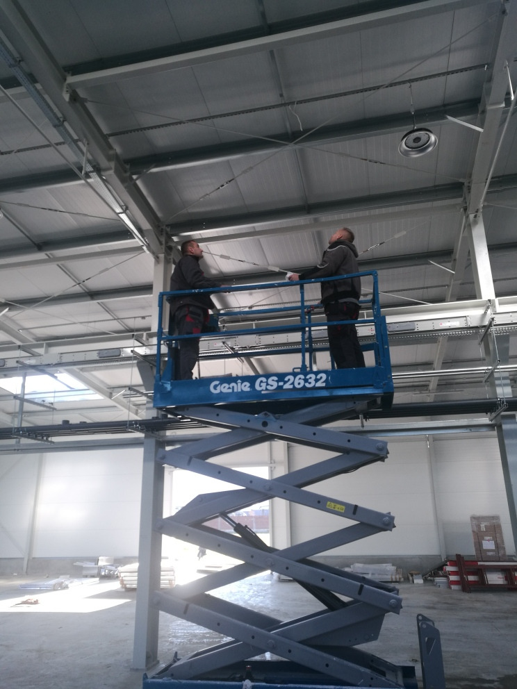 Dwóch mężczyzn na podnośniku Genie GS-2632 montuje elementy pod sufitem w hali przemysłowej, widoczne przewody i konstrukcja sufitu.