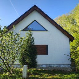 DOMY ZIELONEJ SYNERGII SPÓŁKA Z OGRANICZONĄ ODPOWIEDZIALNOŚCIĄ - Biały dom szkieletowy z trójkątnym oknem i drewnianą elewacją, otoczony zielenią. Ujęcie z poziomu gruntu, w słoneczny dzień.