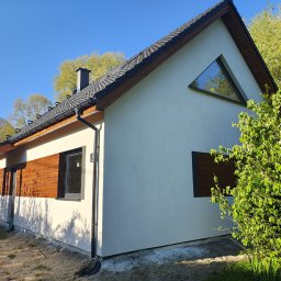 DOMY ZIELONEJ SYNERGII SPÓŁKA Z OGRANICZONĄ ODPOWIEDZIALNOŚCIĄ - Nowoczesny dom z białym tynkiem, drewnianą elewacją i trójkątnym oknem szczytowym. Widoczne orynnowanie i fragment ogrodu z kwitnącym krzewem.