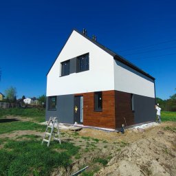 DOMY ZIELONEJ SYNERGII SPÓŁKA Z OGRANICZONĄ ODPOWIEDZIALNOŚCIĄ - Nowoczesny dom z drewnianą elewacją i białymi ścianami, malowany przez pracownika w białym kombinezonie. Widoczne rusztowanie i teren budowy.