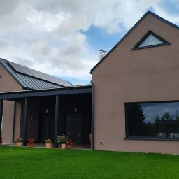 DOMY ZIELONEJ SYNERGII SPÓŁKA Z OGRANICZONĄ ODPOWIEDZIALNOŚCIĄ - Nowoczesny dom modułowy z brązową elewacją, dużymi oknami i panelem słonecznym na dachu. Widok z zewnątrz na zadbany trawnik i elementy architektury.