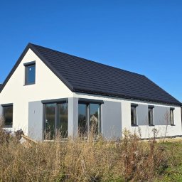 DOMY ZIELONEJ SYNERGII SPÓŁKA Z OGRANICZONĄ ODPOWIEDZIALNOŚCIĄ - Nowoczesny dom z ciemną dachówką i szarymi akcentami, otoczony wysoką trawą. Widoczny drugi dom w tle, błękitne niebo. Ujęcie w słoneczny dzień.