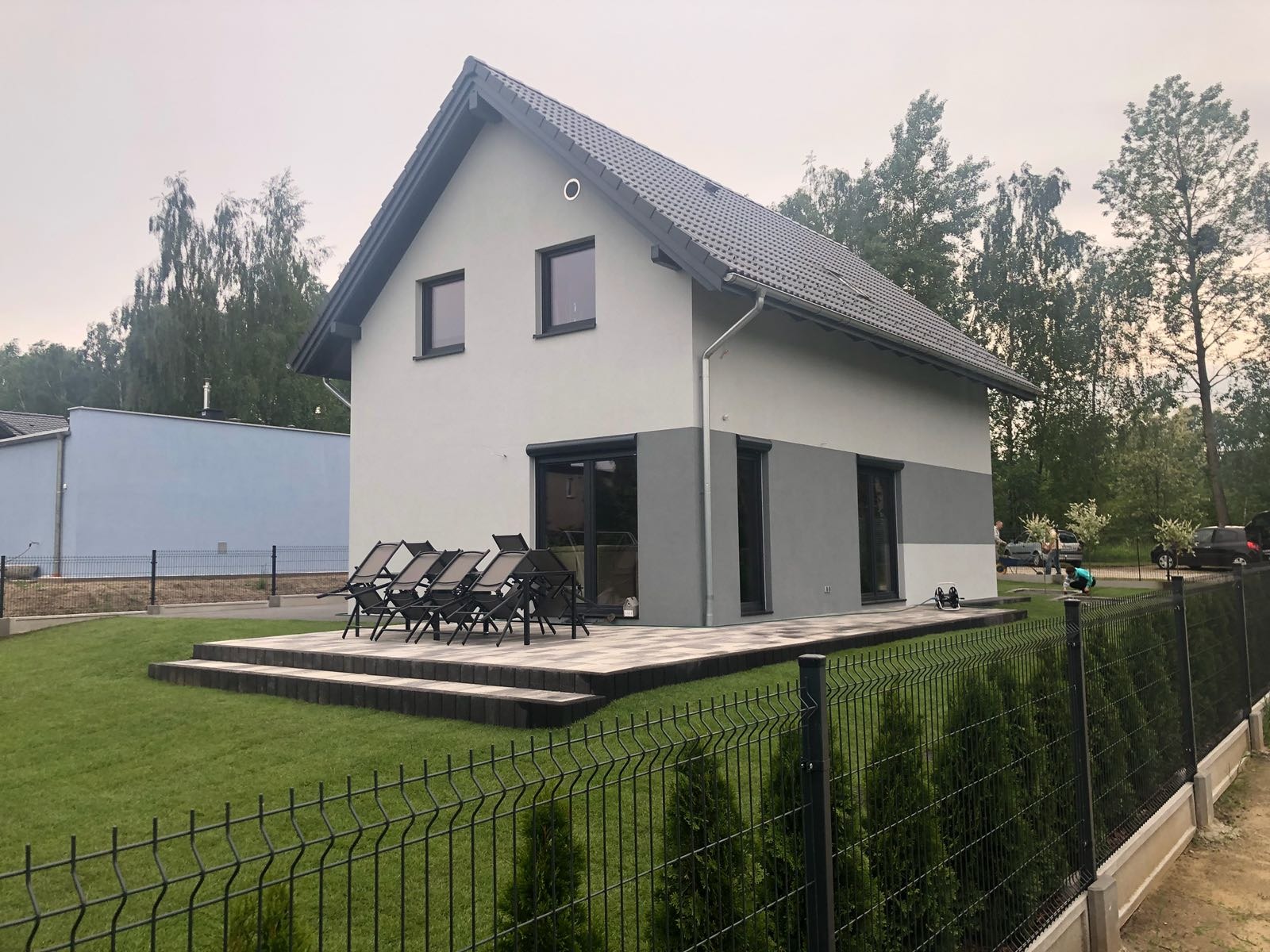 Nowoczesny dom z szarą elewacją, ciemną dachówką i tarasem z meblami ogrodowymi, otoczony zielonym trawnikiem i ogrodzeniem z siatki.
