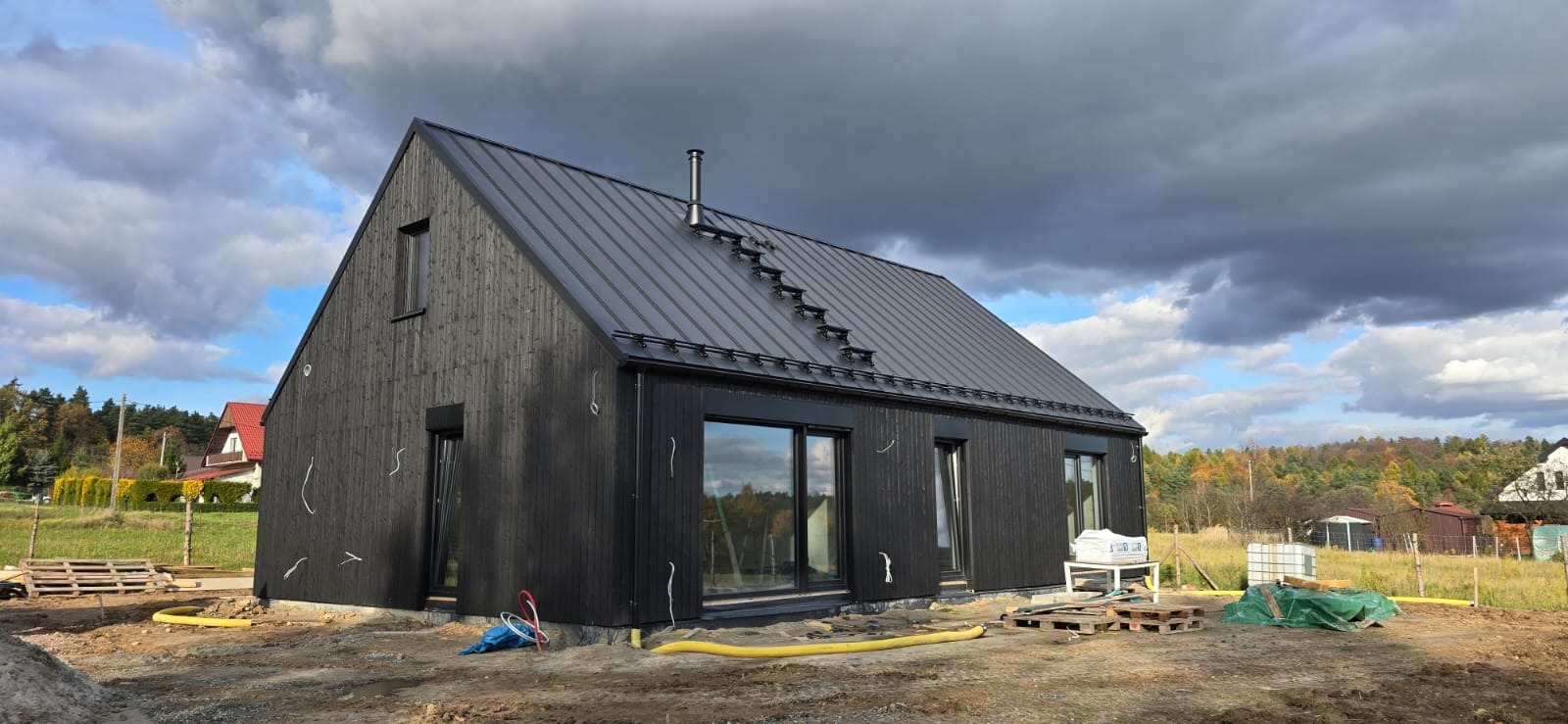 Nowoczesny, czarny dom z drewnianą elewacją i metalowym dachem na etapie wykończenia. Widoczne okna, rury i palety. Tło stanowią drzewa i pochmurne niebo.