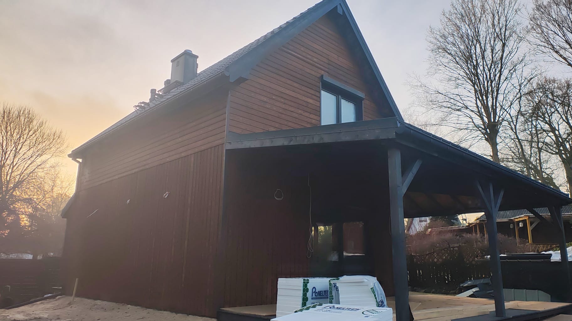 Ciemnobrązowy dom z drewnianą elewacją i czarnym dachem, widok z boku, z widocznymi materiałami budowlanymi u podstawy. Częściowo zachmurzone niebo.