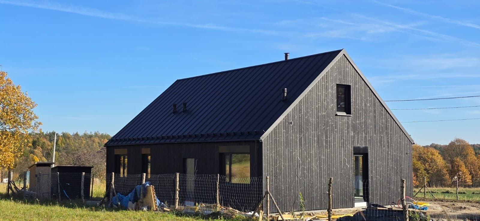 Nowoczesny dom z ciemnego drewna z metalowym dachem, otoczony zielenią i jesiennymi drzewami, widoczny z zewnątrz, w słoneczny dzień.