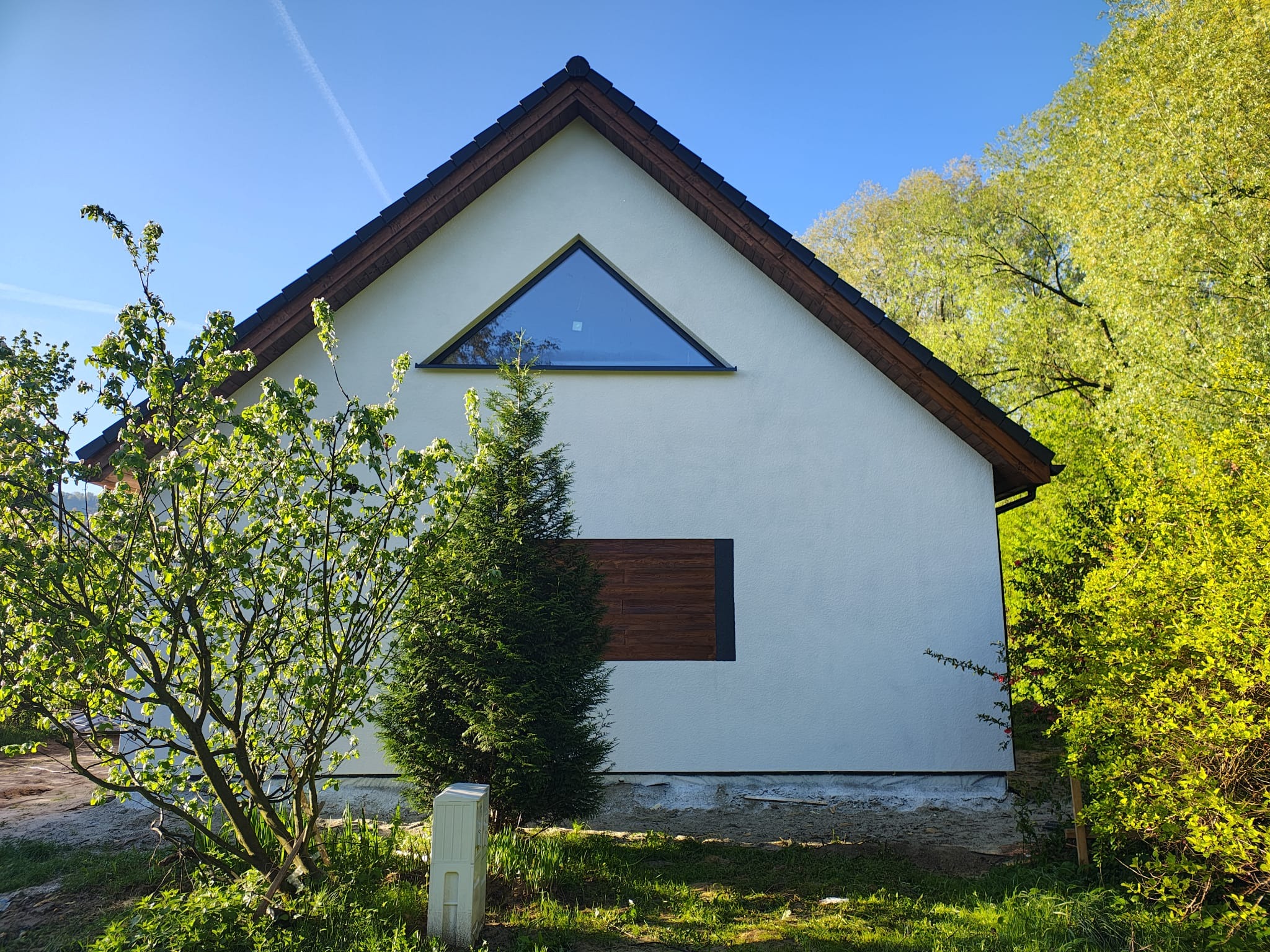Biały dom szkieletowy z trójkątnym oknem i drewnianą elewacją, otoczony zielenią. Ujęcie z poziomu gruntu, w słoneczny dzień.