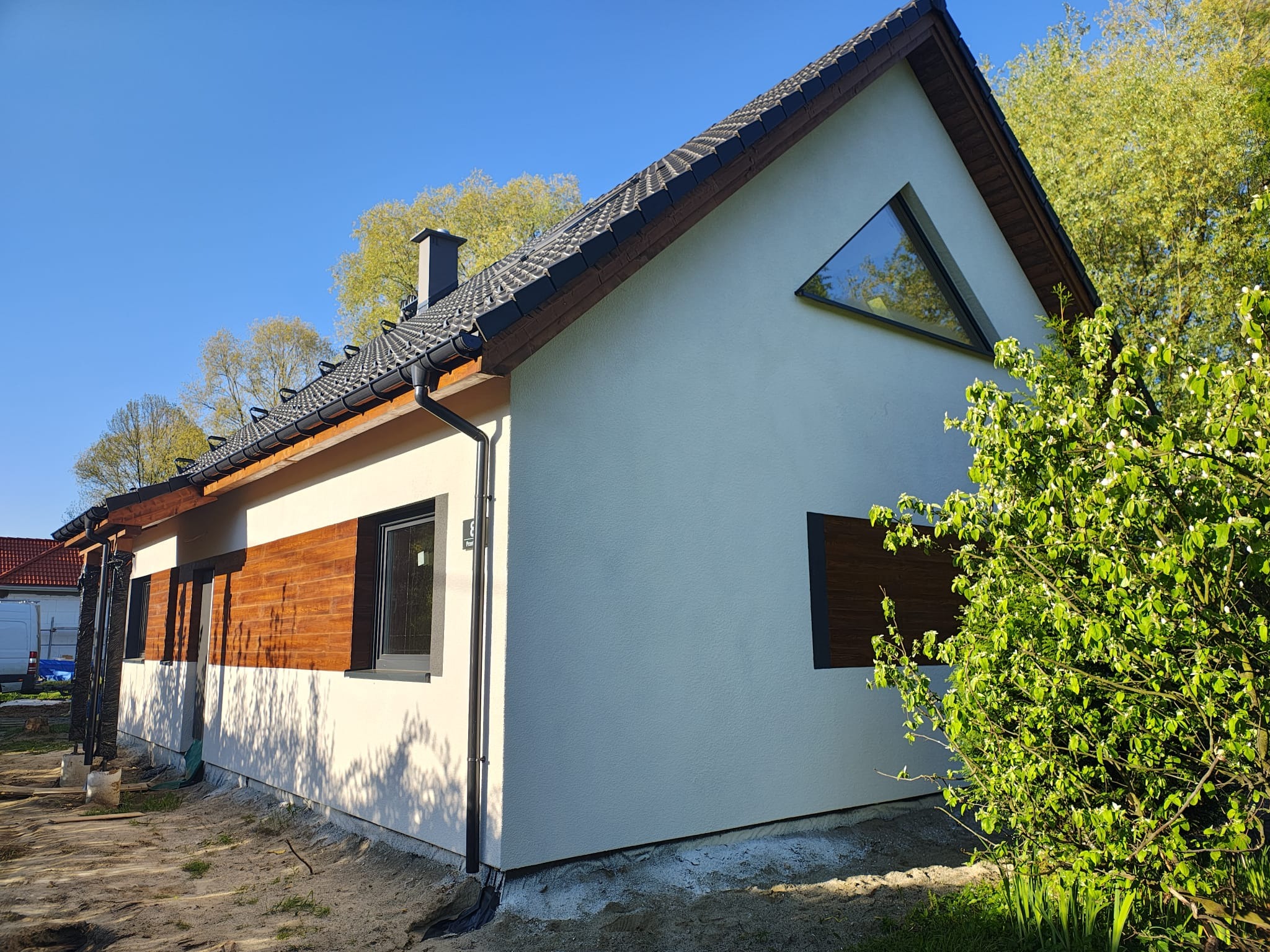 Nowoczesny dom z białym tynkiem, drewnianą elewacją i trójkątnym oknem szczytowym. Widoczne orynnowanie i fragment ogrodu z kwitnącym krzewem.