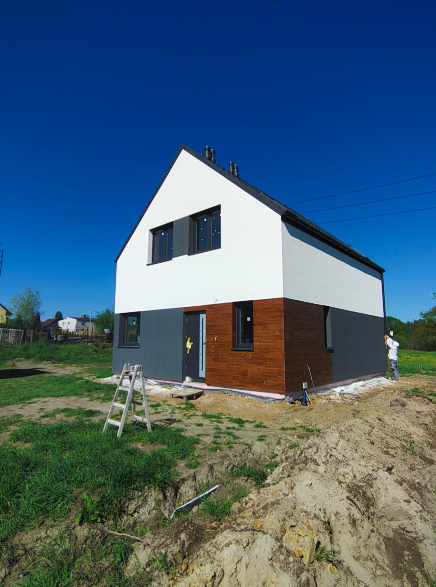 Nowoczesny dom z drewnianą elewacją i białymi ścianami, malowany przez pracownika w białym kombinezonie. Widoczne rusztowanie i teren budowy.