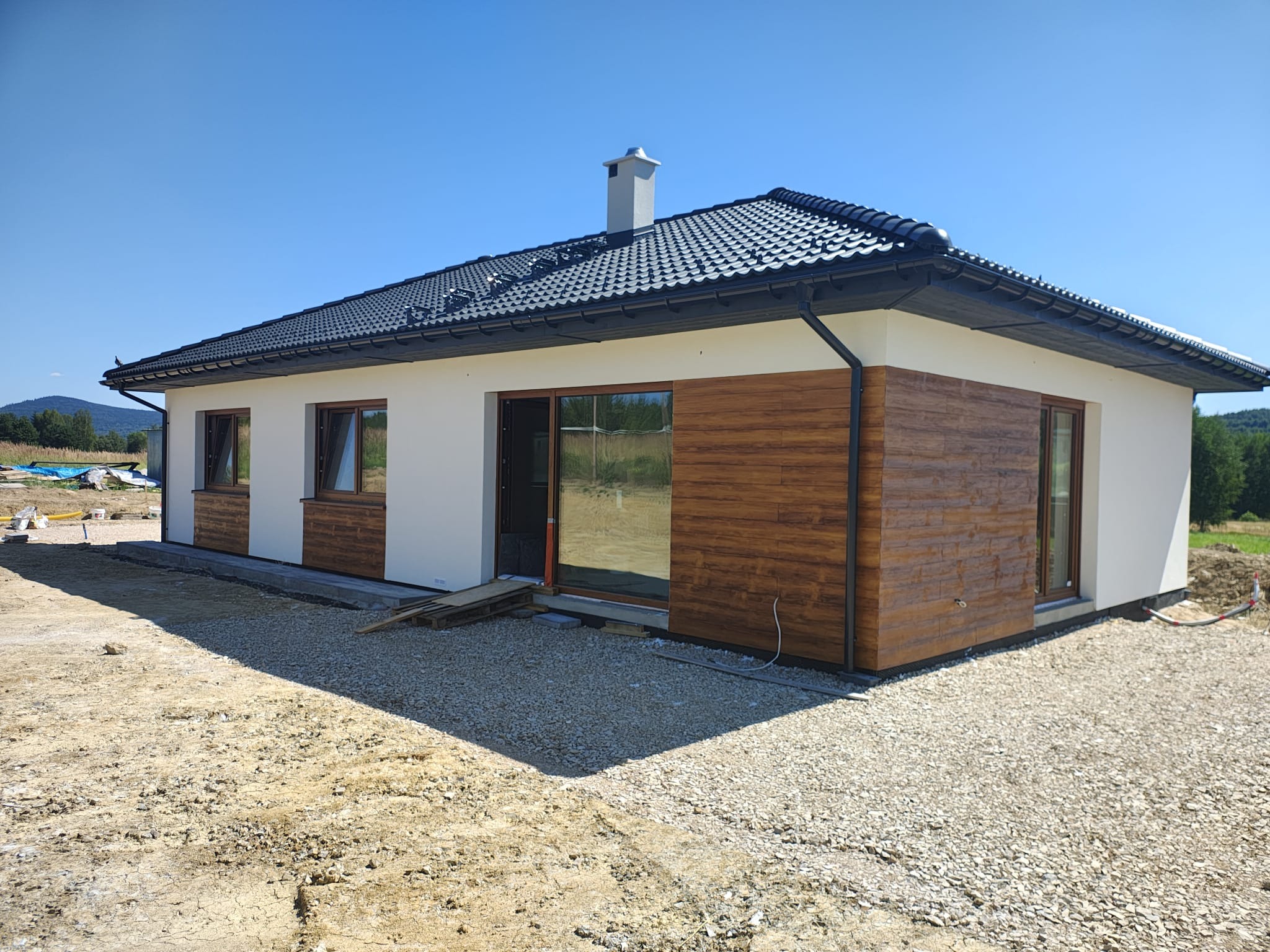 Parterowy dom z ciemną dachówką i drewnianą elewacją, jasne ściany, duże okna, w tle zielone wzgórza pod błękitnym niebem. Dom w trakcie wykańczania.