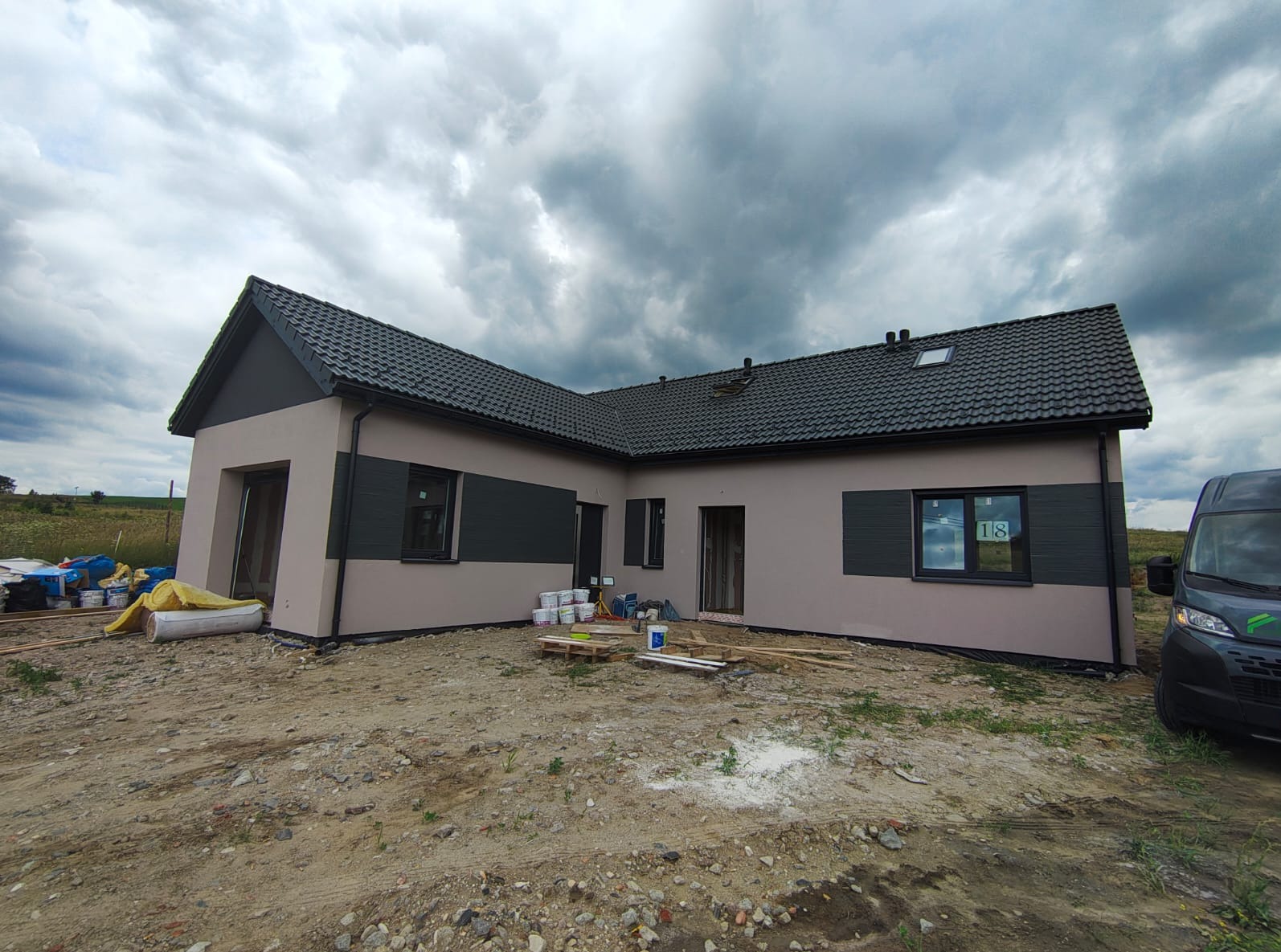 Parterowy dom z ciemną dachówką i elementami elewacji w kolorze grafitowym, widoczny z zewnątrz, na tle pochmurnego nieba. Częściowo zagospodarowany teren.