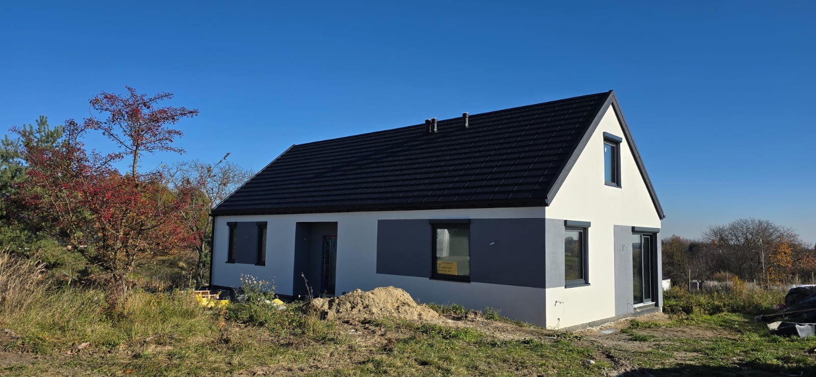 Gotowy, parterowy dom z dwuspadowym dachem i szarą elewacją, otoczony zielenią i drzewami pod błękitnym niebem. Widoczny fragment placu budowy.