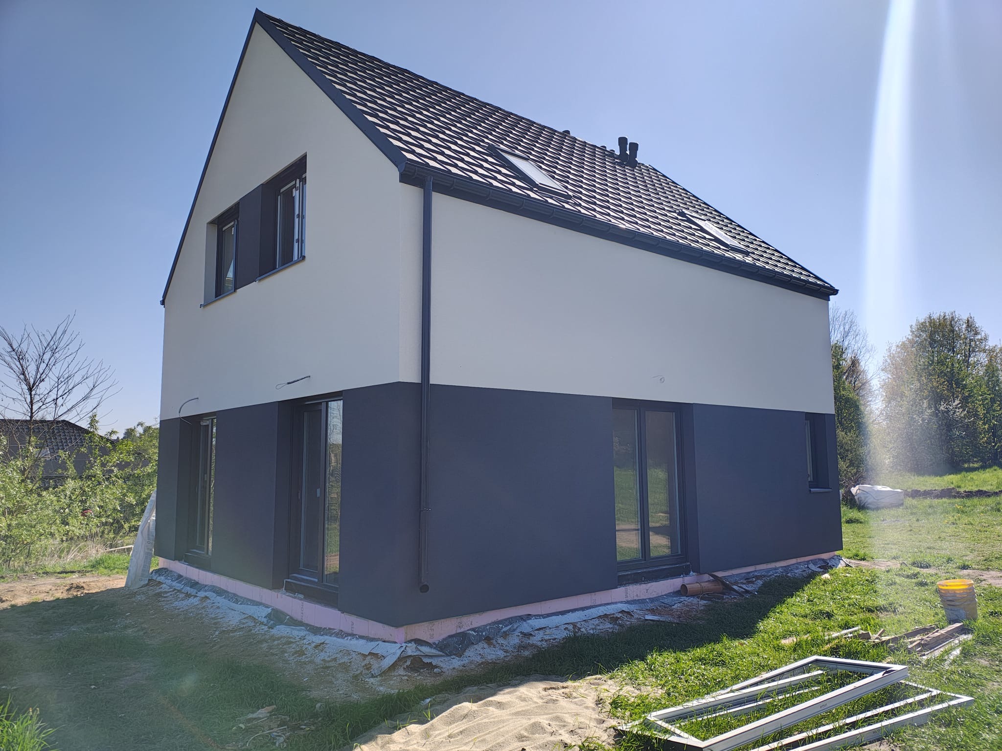 Dwukondygnacyjny dom z ciemnoszarą dolną i jasną górną elewacją, czarnym dachem i oknami, otoczony zielenią i piaskiem, w słoneczny dzień.