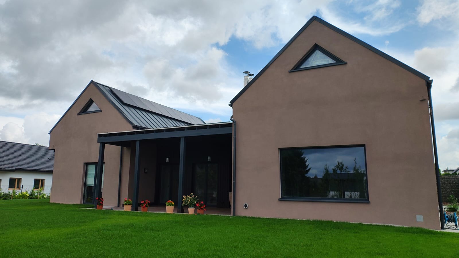 Nowoczesny dom modułowy z brązową elewacją, dużymi oknami i panelem słonecznym na dachu. Widok z zewnątrz na zadbany trawnik i elementy architektury.