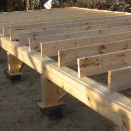 Konstrukcja podłogi z drewnianych belek na fundamentach z izolacją, widoczne sznurki traserskie, przygotowana pod dalsze etapy budowy.