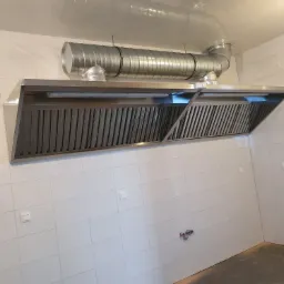 Profesjonalny, stalowy okap przemysłowy z widocznymi rurami wentylacyjnymi zamontowany w pomieszczeniu z białymi, kwadratowymi płytkami.