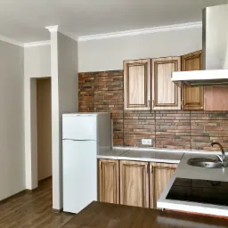 Wykończona kuchnia z drewnianymi szafkami, ceglaną mozaiką nad blatem i białym sufitem z dekoracyjną listwą, widok na otwarte przejście do innego pomieszczenia.