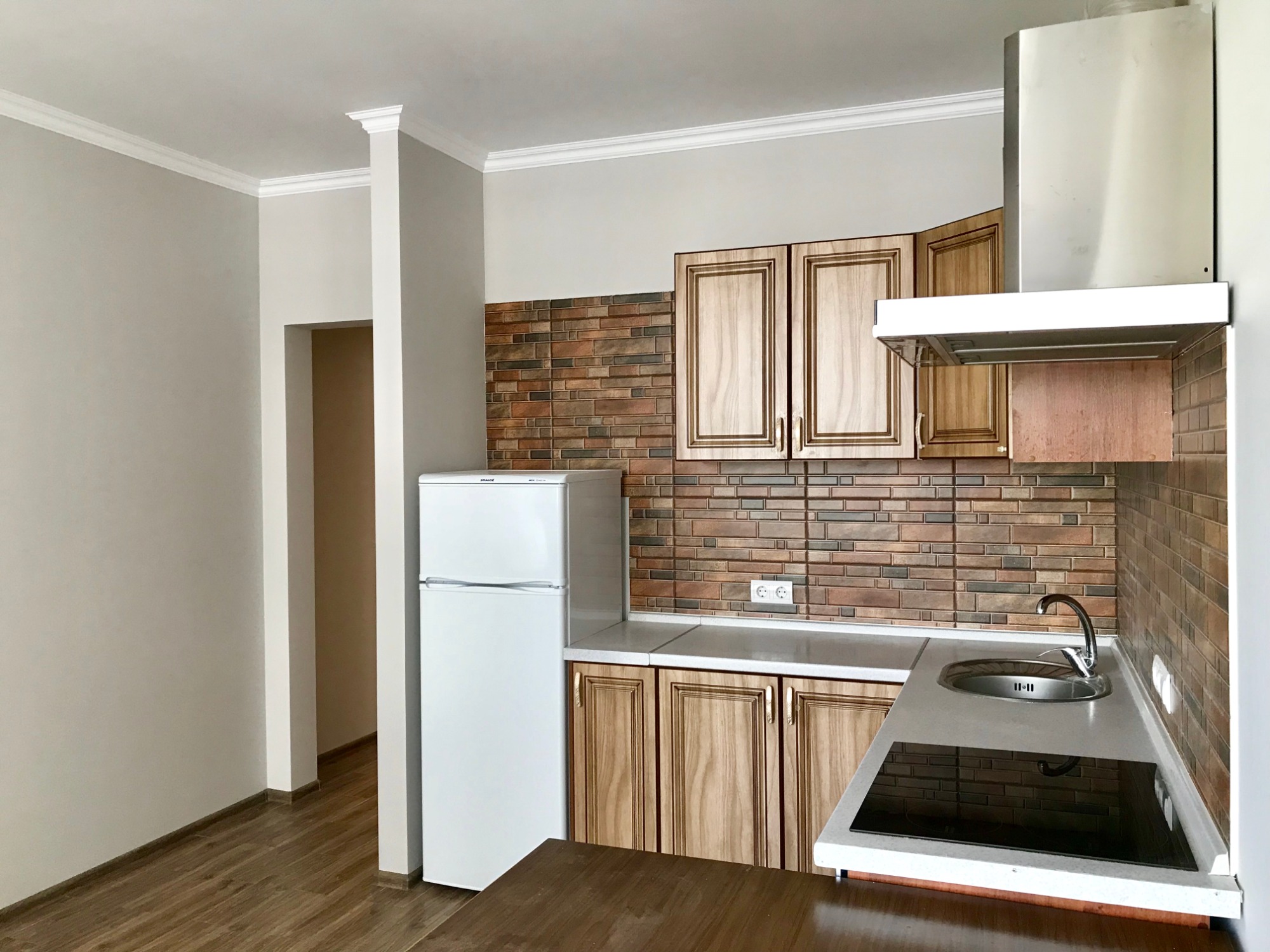 Wykończona kuchnia z drewnianymi szafkami, ceglaną mozaiką nad blatem i białym sufitem z dekoracyjną listwą, widok na otwarte przejście do innego pomieszczenia.