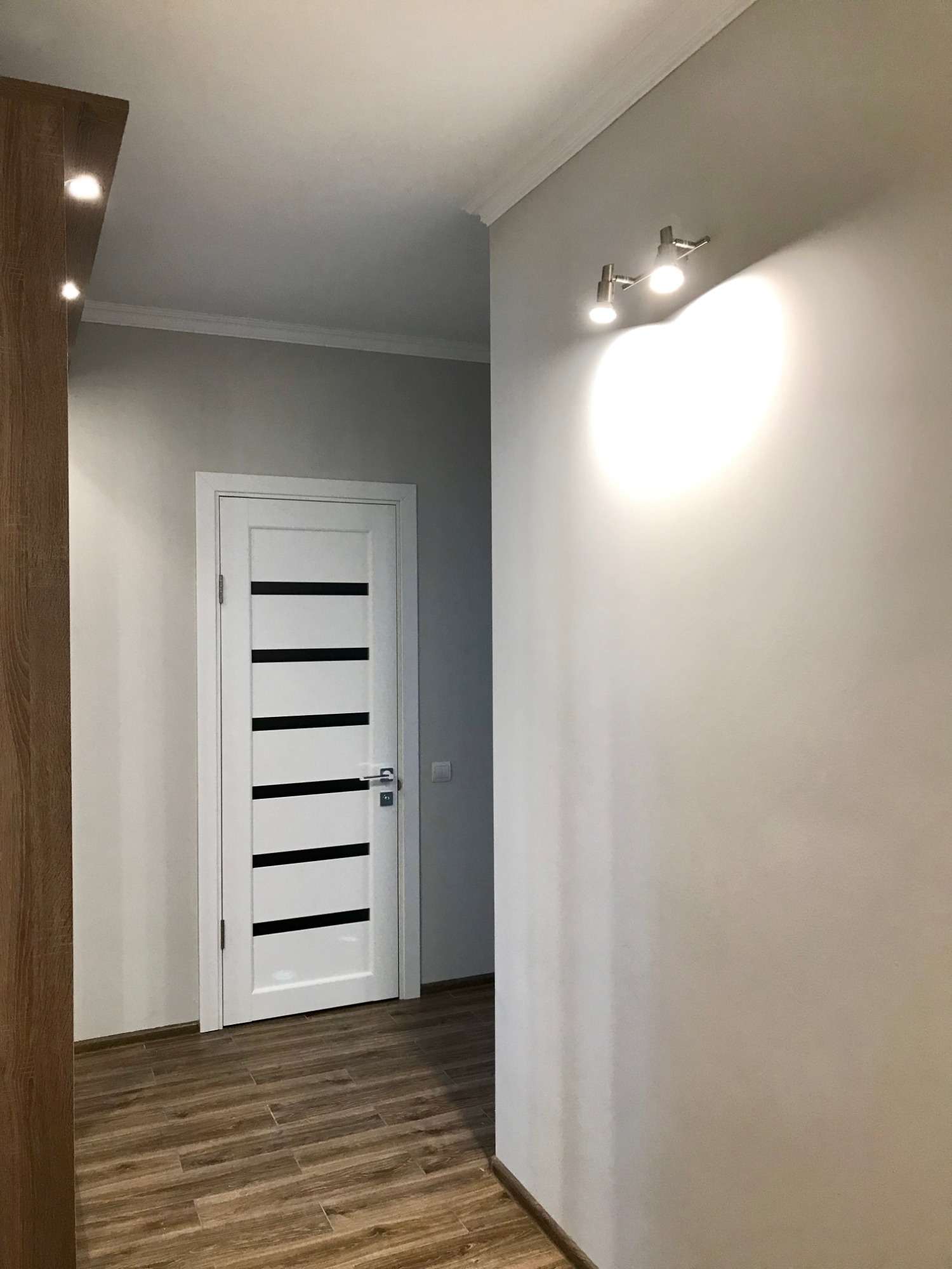 Korytarz po remoncie: białe ściany, drewnopodobna podłoga, białe drzwi z czarnymi paskami, oświetlenie punktowe na ścianie.