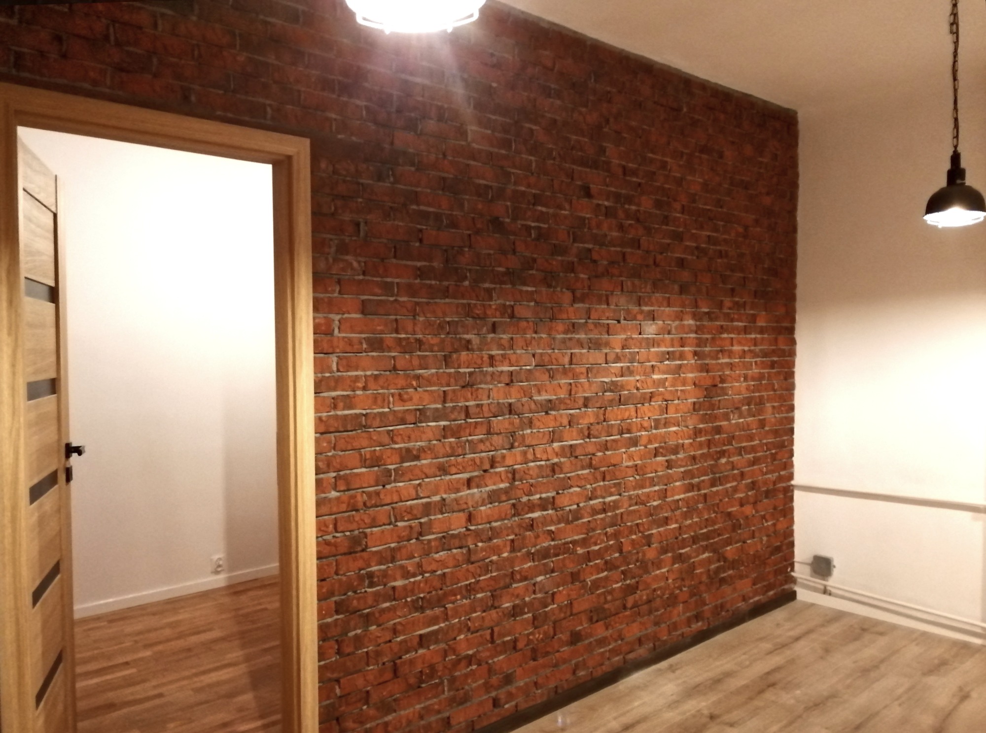 Ściana z czerwonej cegły w pokoju po remoncie w Legionowie, z widocznymi śladami fugi i oświetleniem górnym.