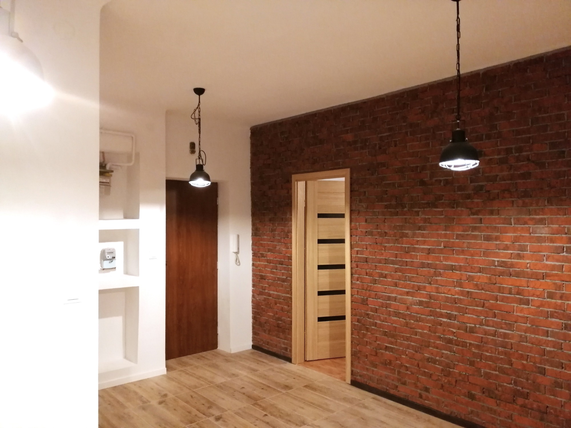 Wnętrze mieszkania z ceglaną ścianą, drewnianą podłogą, dwiema wiszącymi lampami w stylu industrialnym, białą ścianą z wnękami i drewnianymi drzwiami.