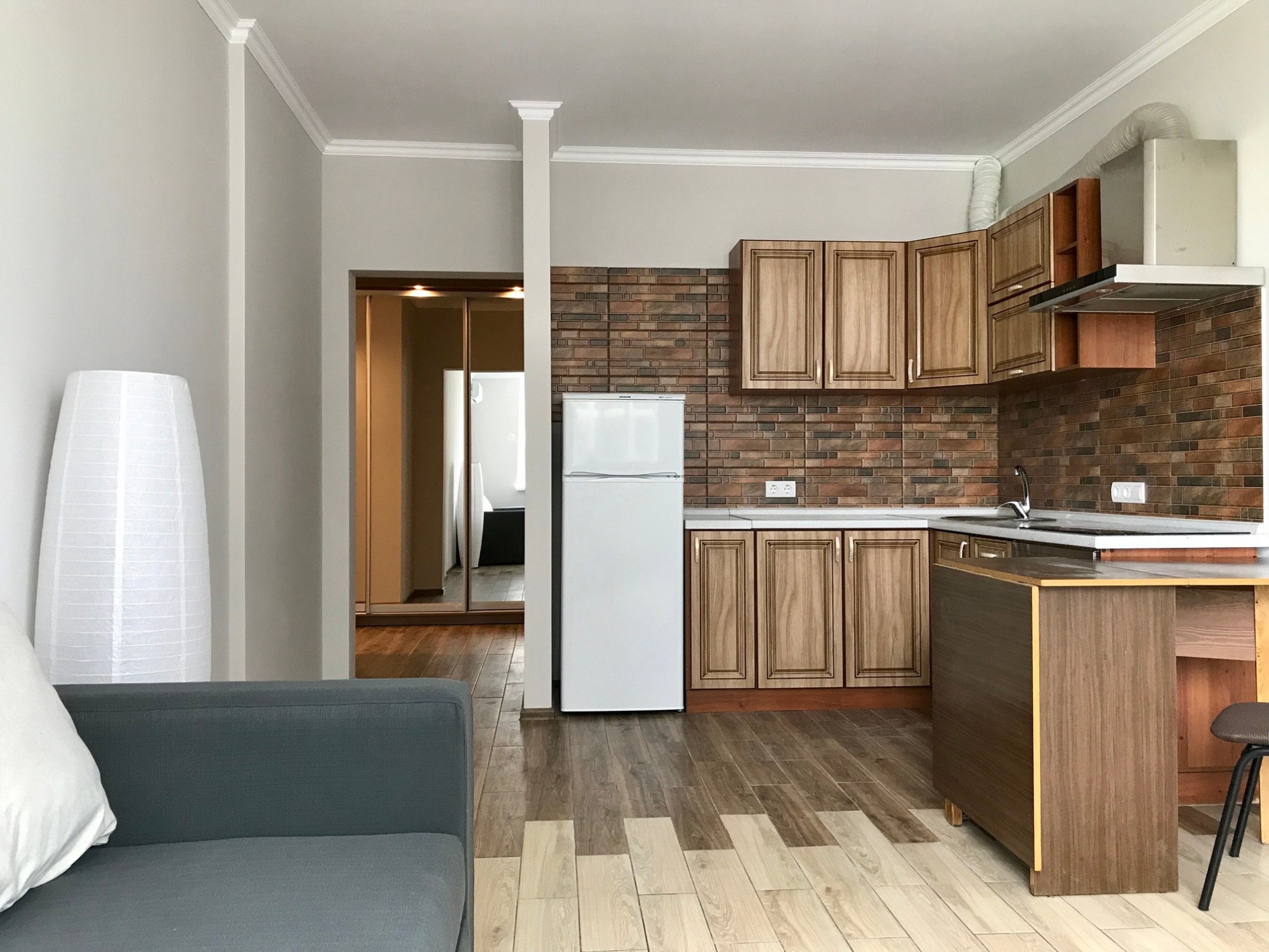 Jasne mieszkanie po remoncie: widok na kuchnię z brązowymi szafkami i ceglaną ścianą, białą lodówką oraz salon z szarą sofą i lampą w tle.