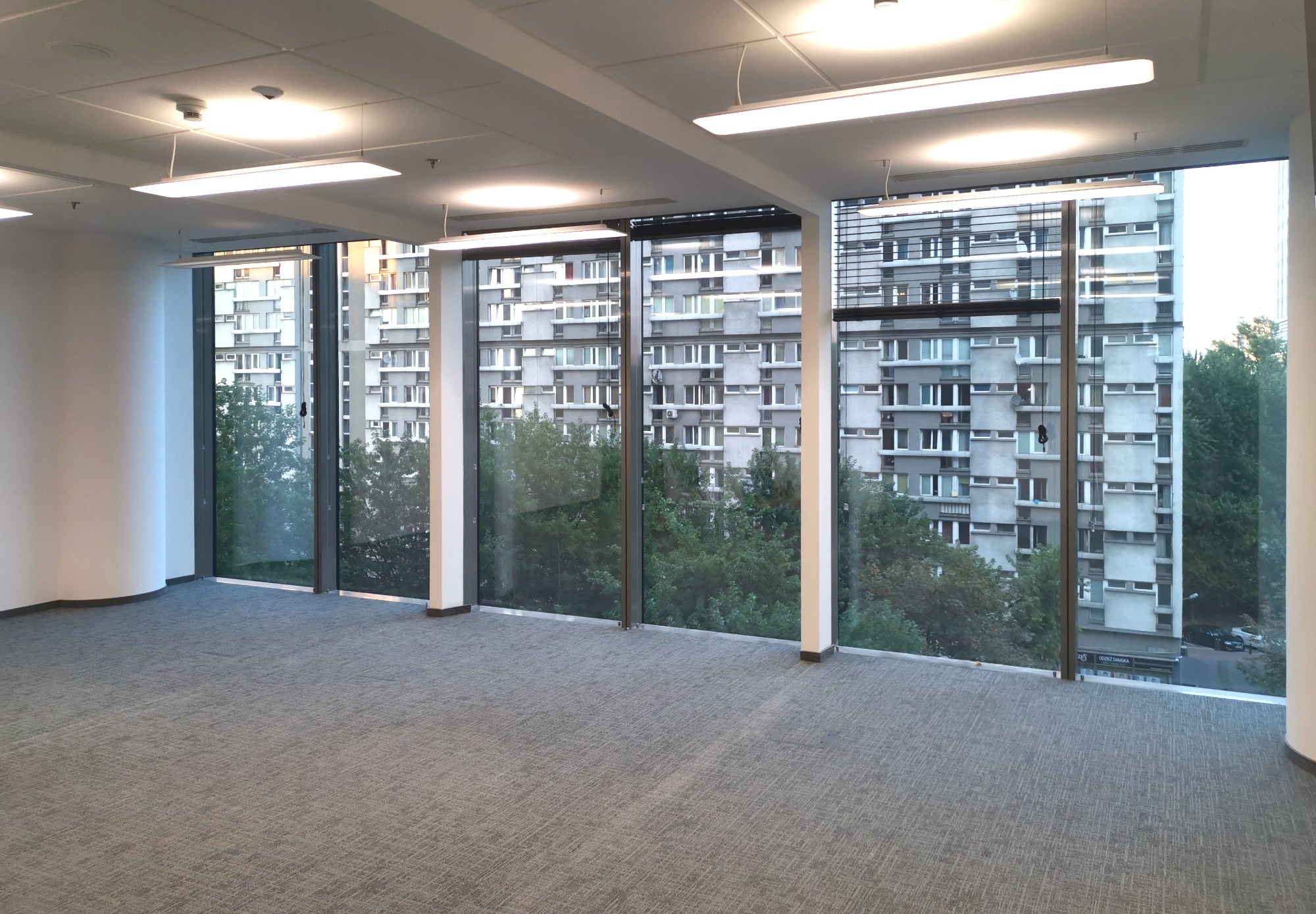 Puste, jasne biuro z szarą wykładziną dywanową i dużymi oknami z widokiem na blok mieszkalny i zieleń, oświetlone podwieszanymi lampami.